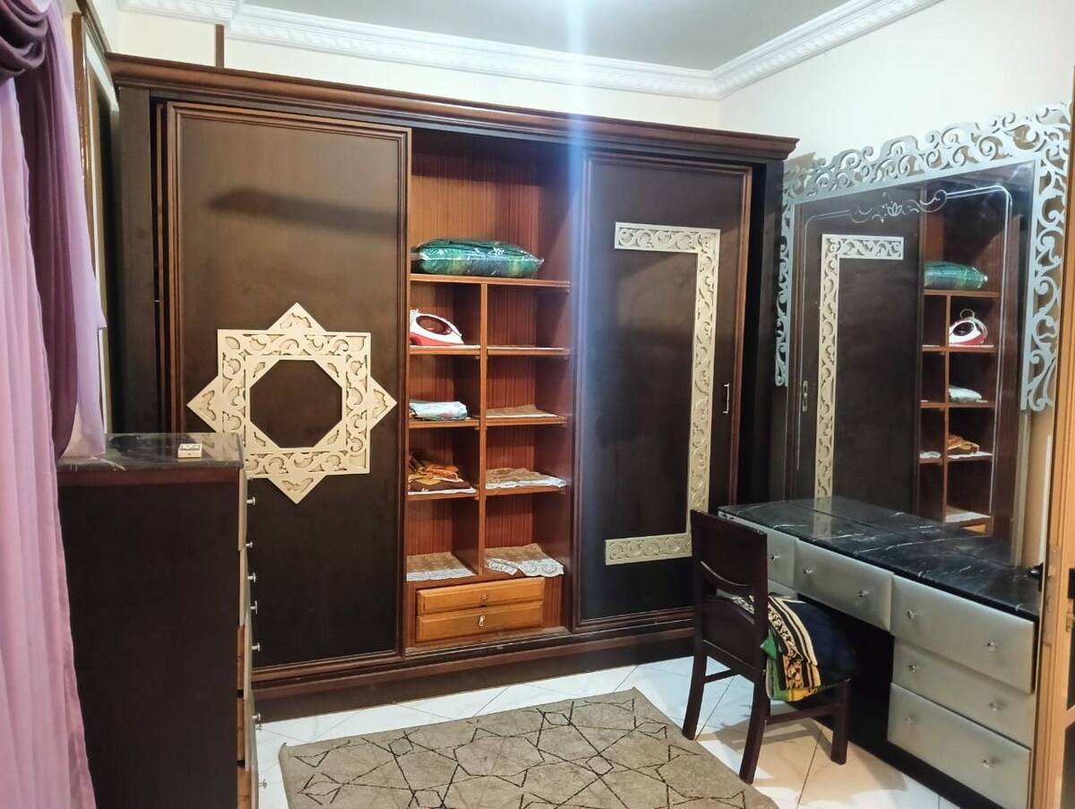 A wooden wardrobe with intricate design details stands against the wall, offering open shelving and a drawer. A modern black vanity table is positioned nearby, accompanied by a mirror with decorative framing. Soft lighting enhances the space, creating a functional and stylish atmosphere.