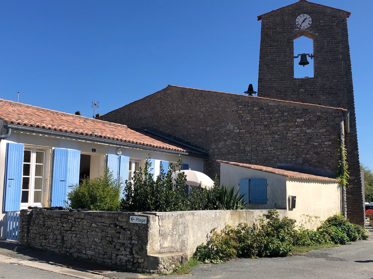 A charming stone exterior with light blue shutters faces the street, complemented by a neatly kept garden. Adjacent, a tall clock tower with a bell is visible against a clear blue sky, enhancing the tranquil village setting.