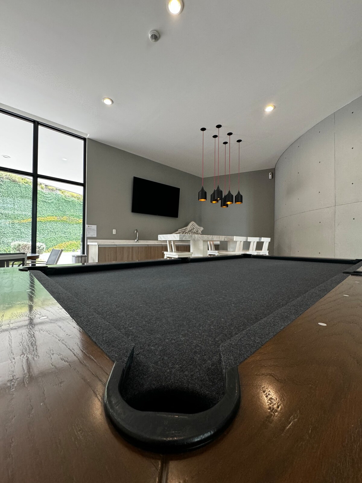 A pool table is featured in the foreground, with a sleek wooden edge and a dark playing surface. In the background, a light-filled area is visible, showcasing a modern seating arrangement and a television mounted on the wall. Decorative pendant lights hang above.