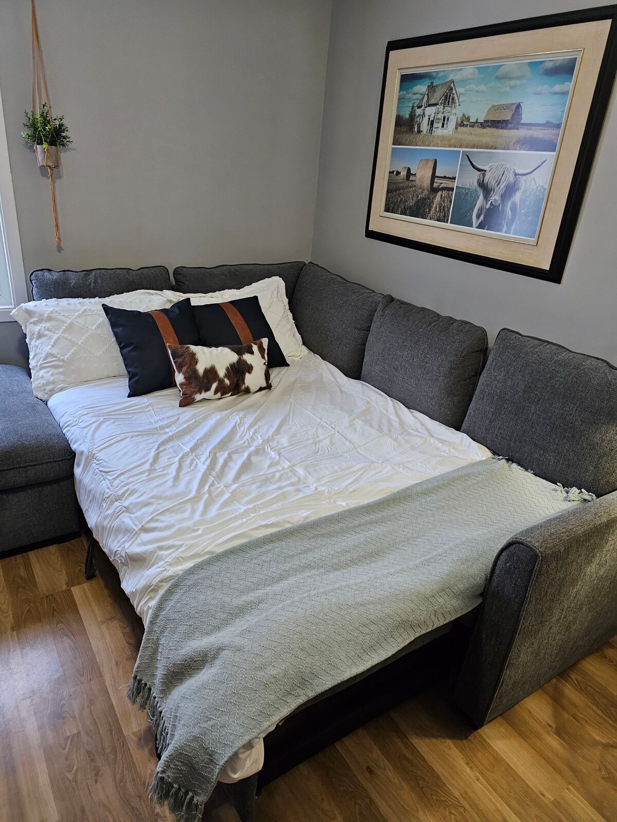 A cozy living area features a gray sectional sofa, with a pull-out bed prepared with a white quilt and decorative pillows. A soft throw blanket is draped over the arm, and an art piece depicting rural scenery hangs above the seating.
