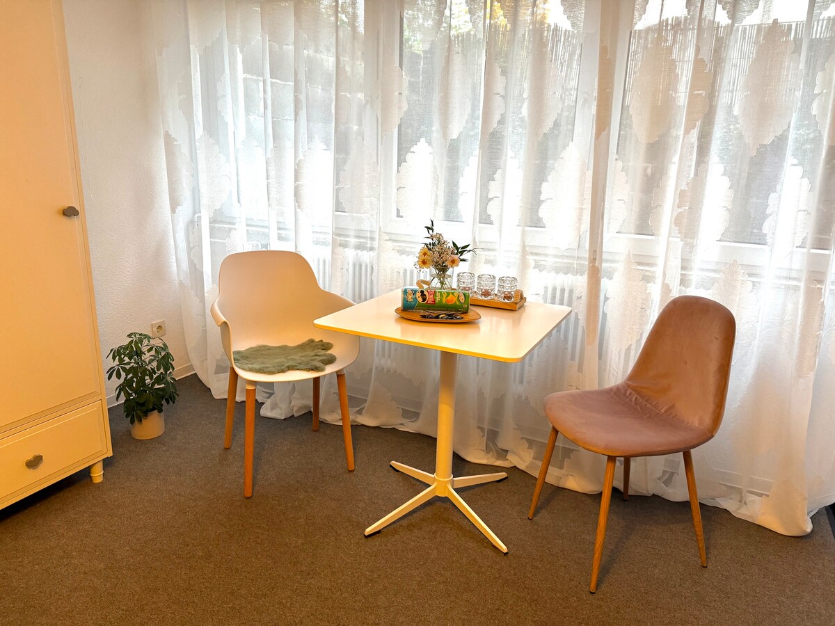 A dining area features a small round table positioned between two stylish chairs, one with a light hue and the other in a muted tone. Natural light streams through sheer curtains, creating a bright ambiance. A potted plant adds a touch of greenery to the corner.