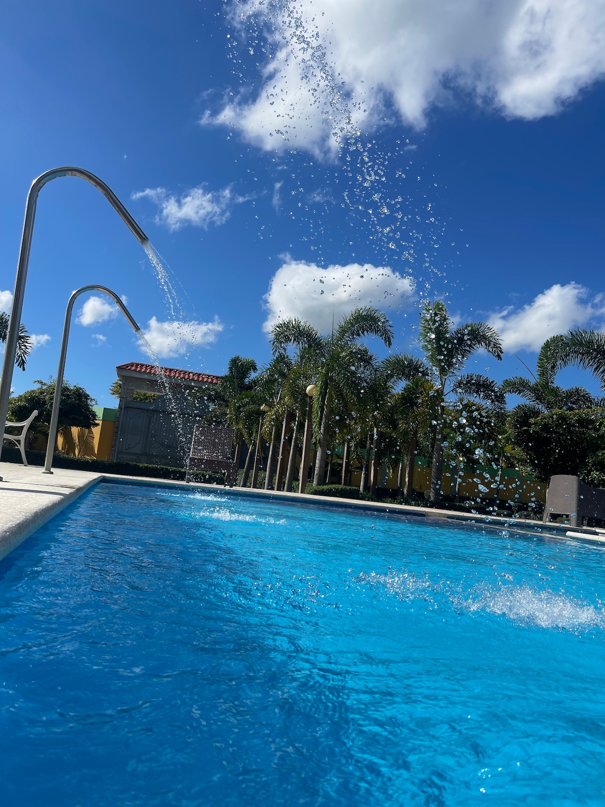 A clear swimming pool is depicted, with water splashing from elevated features on the sides. Lush green palm trees surround the area, standing against a bright blue sky dotted with fluffy white clouds.
