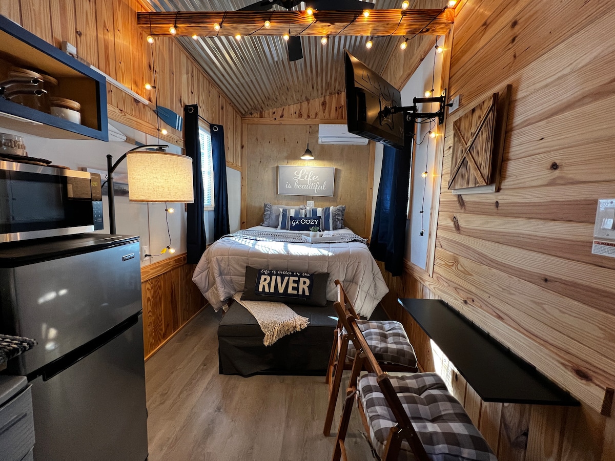 The interior of the tiny home features warm wooden walls and soft lighting. A queen bed is prominently placed, adorned with decorative pillows. A small kitchenette is visible, alongside a seating area with two stools and a table, creating an inviting space.