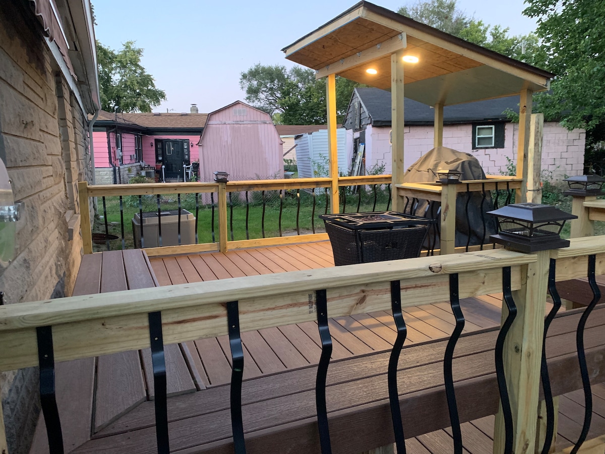A wooden deck is presented with a covered seating area featuring a table and chairs. Two lanterns provide soft illumination, while a grill is positioned nearby. The surrounding yard includes greenery and structures further in the background, creating a spacious outdoor setting.
