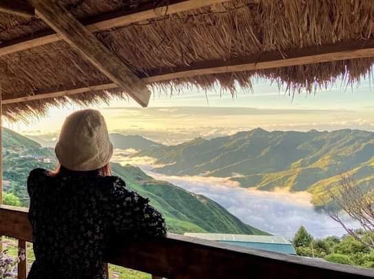 A scenic view from a covered terrace reveals rolling mountains blanketed in mist. A person is seen gazing into the distance, with a straw roof overhead providing shade. The horizon displays a soft blend of colors as the sun rises.