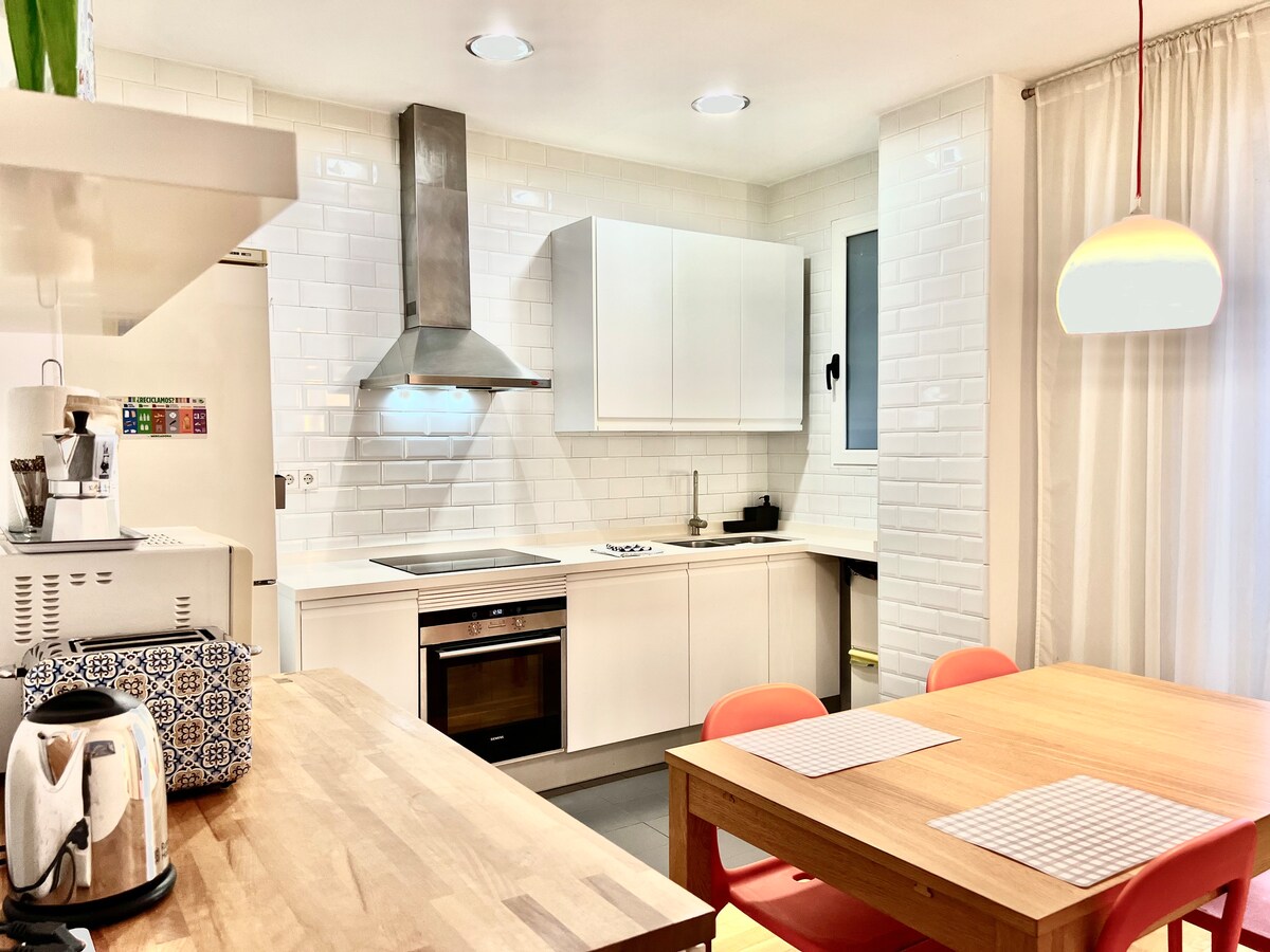 A modern kitchen features sleek white cabinetry and a stainless-steel oven. A wooden table with red chairs is positioned for dining. Natural light enters through a window, enhancing the space, while a minimalist light fixture hangs above the table.