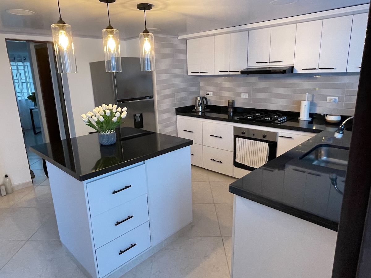 The modern kitchen features sleek white cabinetry and a black countertop, creating a stylish contrast. A central island offers additional counter space and storage. Pendant lights hang above, illuminating the space, while appliances are neatly integrated along the wall, enhancing functionality.