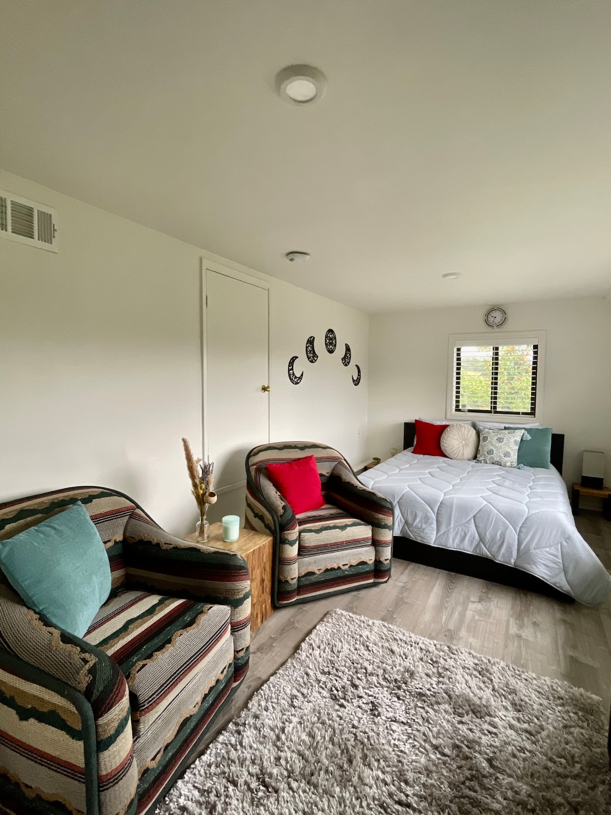 A cozy living space features a queen bed with light bedding, flanked by decorative pillows. Two patterned chairs with red and teal accents complement a soft area rug on the floor. Natural light enters through a window with simple blinds, illuminating the room's neutral decor.