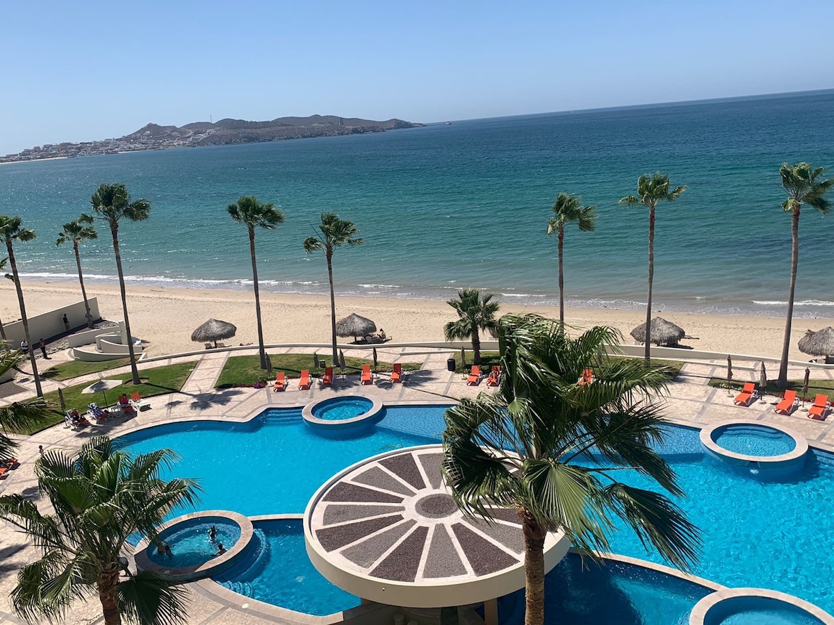 The image presents a view from a higher floor, showcasing a large swimming pool surrounded by palm trees. The pool area features sun loungers and four jacuzzis. Beyond the pool, a sandy beach meets the shimmering blue water of the ocean, enhancing the appealing coastal scenery.