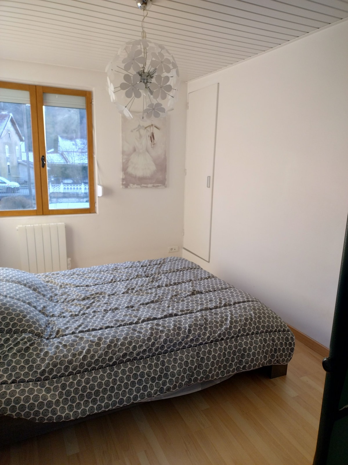 A bedroom features a spacious bed dressed in patterned bedding, positioned near a window that allows natural light to enter. A decorative light fixture hangs from the ceiling, and a wall-mounted artwork adds a touch of style. The floor is covered in light wood.