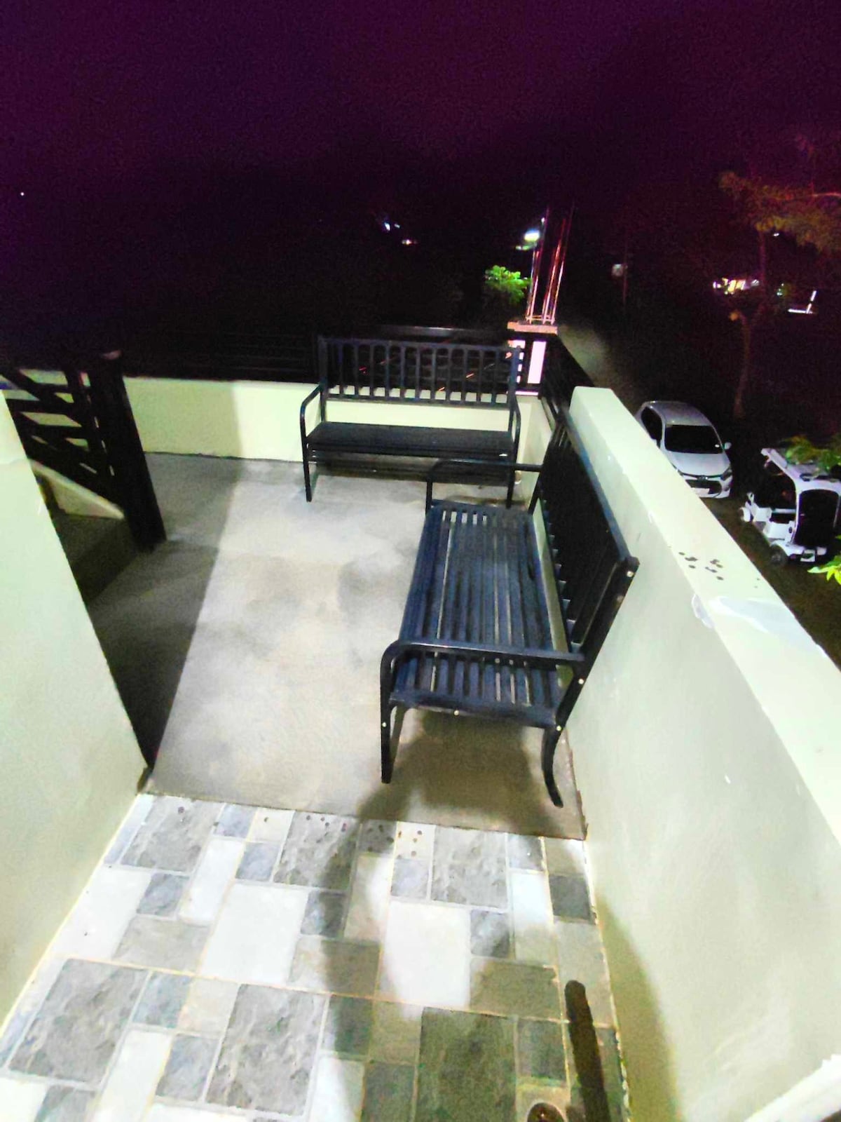 A rear balcony features two black metal benches placed against a low wall. The flooring is comprised of patterned tiles, and the area is illuminated by soft outdoor lighting, creating a relaxed atmosphere suitable for quiet moments or enjoying the surroundings.