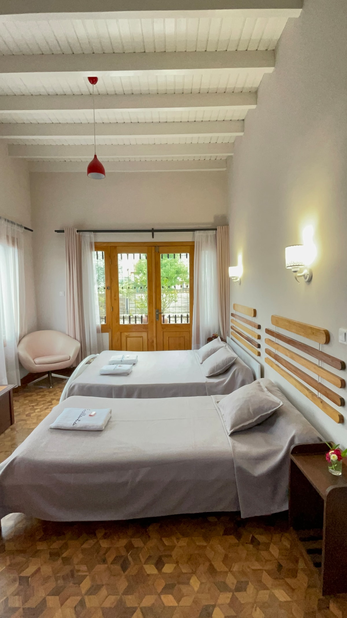 A cozy room with two spacious beds, each adorned with neatly folded towels. Natural light fills the space through large double doors leading to a balcony. Soft-colored walls and wooden accents create a calming environment, complemented by a stylish chair in the corner.