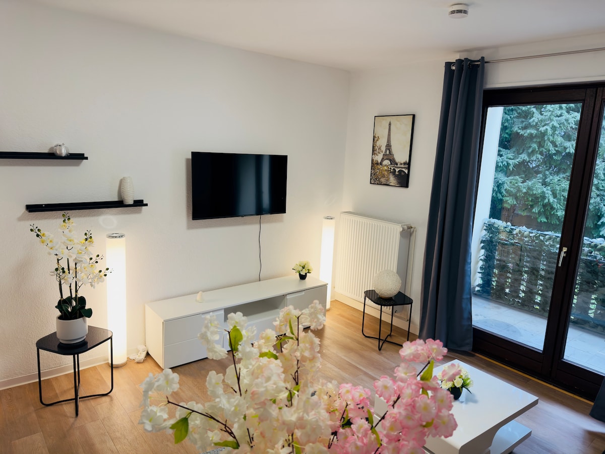 A light-filled living area is showcased with two floor lamps flanking a low white media console. A television is mounted on the white wall. Elegant flower arrangements are positioned on tables, adding a touch of nature, while a door opens to a private outdoor space.