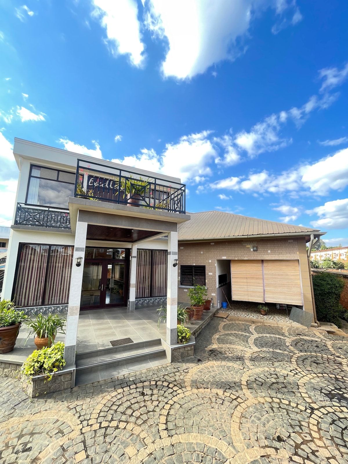 A two-story building is presented with a welcoming entrance featuring a covered porch and decorative plants. A balcony is visible on the upper level, offering a view of the sky. The cobblestone driveway leads to a garage with a wooden door.