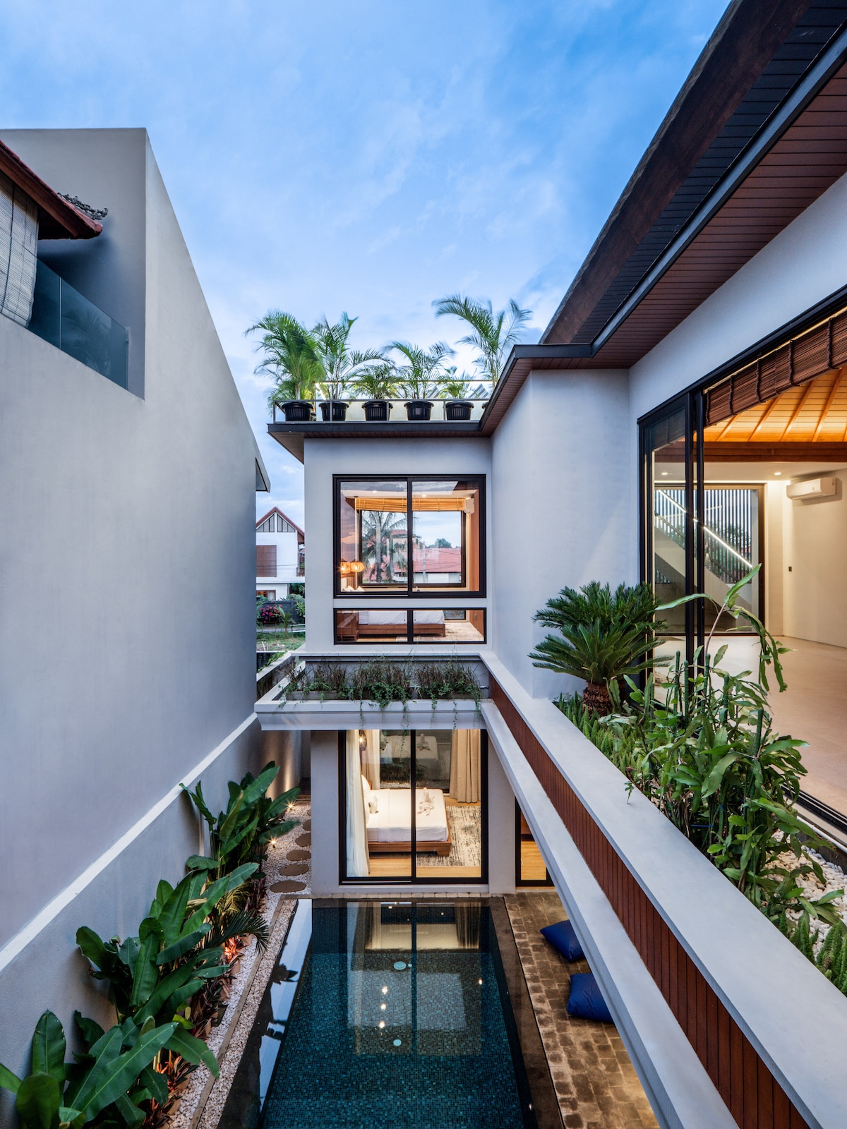 An airy courtyard showcases a clear swimming pool surrounded by lush greenery, with tall palm trees visible in the background. Large glass windows provide a glimpse into the interior spaces, merging natural light with an open, welcoming environment.