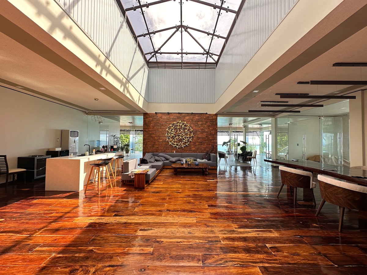 A spacious living area features a high ceiling with large skylights, allowing for abundant natural light. A wooden floor adds warmth to the space. Modern furniture includes a gray sectional sofa and a stylish dining arrangement, complemented by decorative elements on the walls.