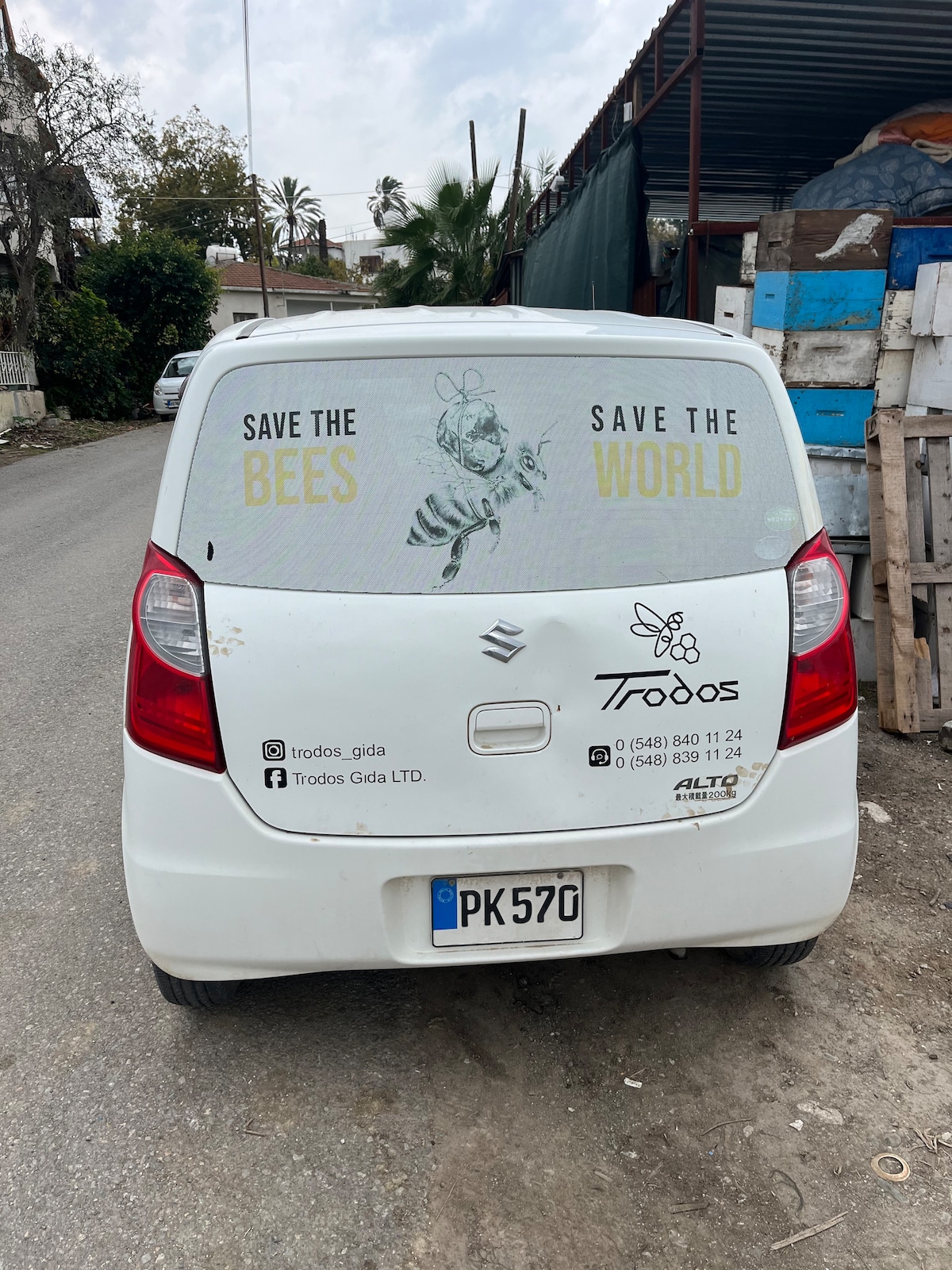 A white vehicle is parked on a narrow street, featuring a window decal with the message 'Save the Bees, Save the World.' The van is surrounded by rustic building materials and vegetation, indicative of the local environment.