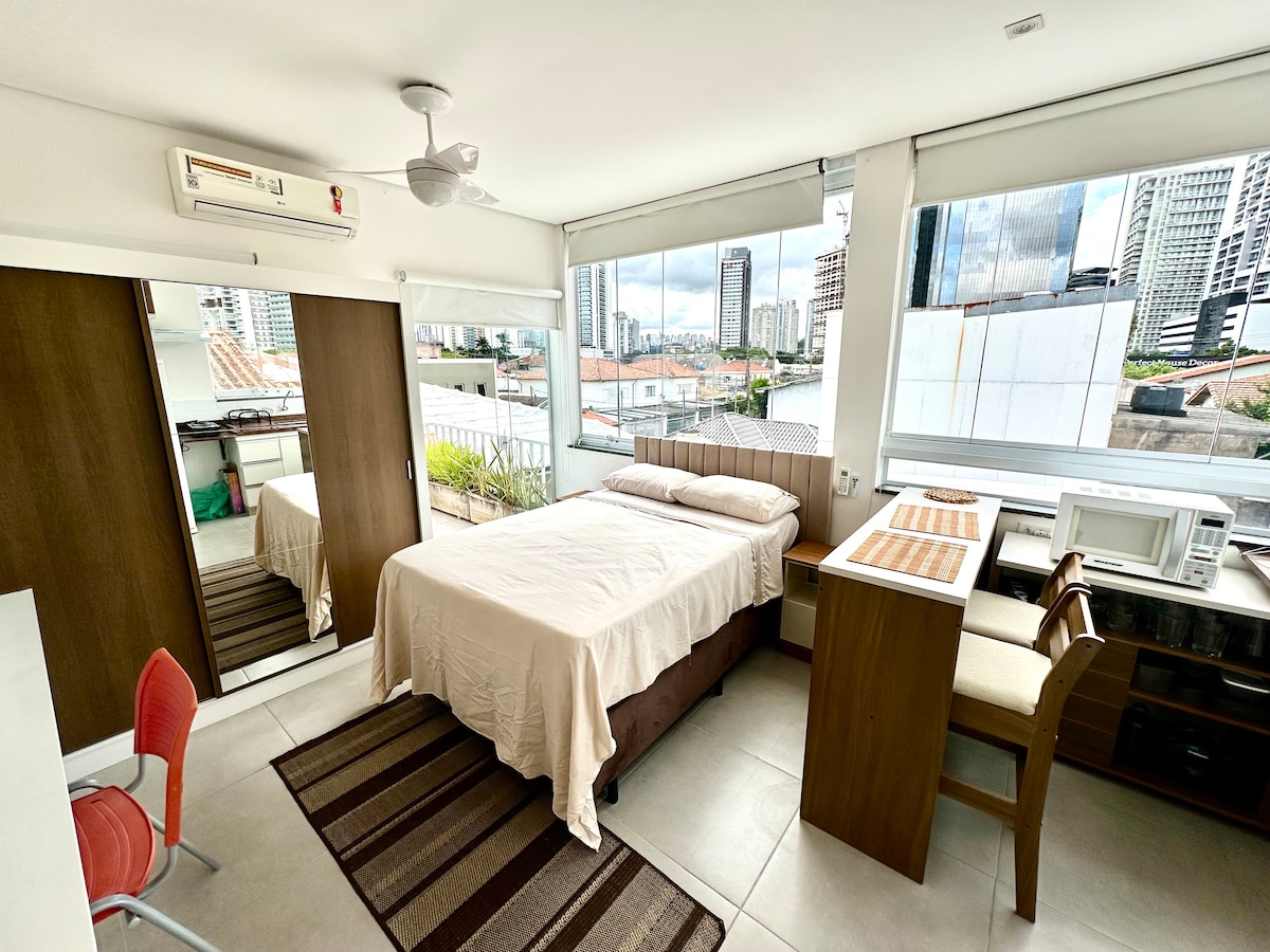 A well-lit studio features a spacious bed with a light-colored coverlet. A large mirror reflects the room, which includes a desk and a microwave. Floor-to-ceiling windows offer a view of the cityscape, while a fan is mounted on the ceiling.