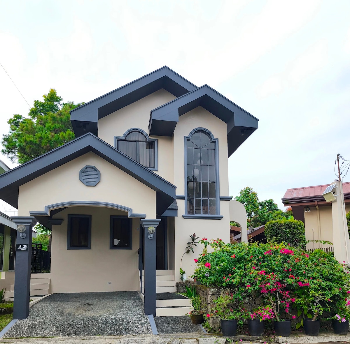The two-story house features a modern design with contrasting colors and arched windows. It is surrounded by vibrant potted plants and shrubs, adding color to the front entrance. A spacious driveway is visible, providing easy access to the home.