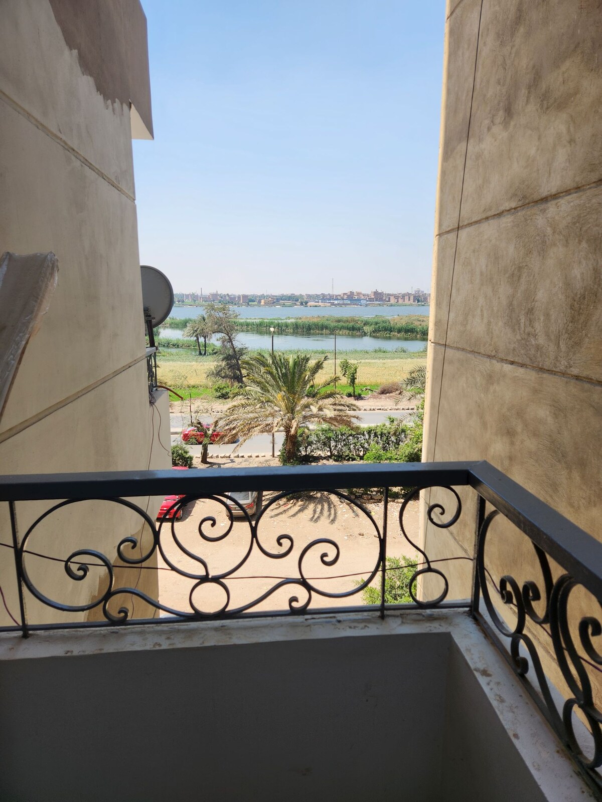 A balcony view overlooking the Nile River is framed by ornamental railing. Lush greenery and palm trees line the riverbank, with urban buildings visible in the distance under a clear sky.