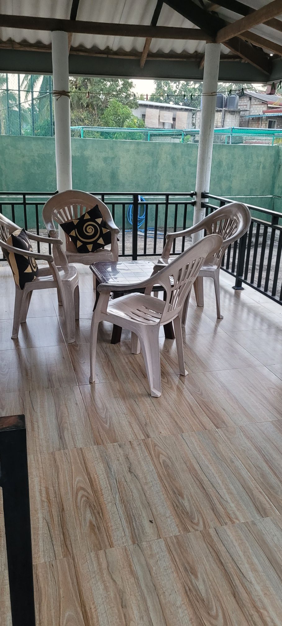 A covered outdoor seating area is presented, featuring four plastic chairs arranged around a small table. The wooden floor complements the neutral tones of the furniture, while green fencing and trees are visible in the background, creating a peaceful ambiance.