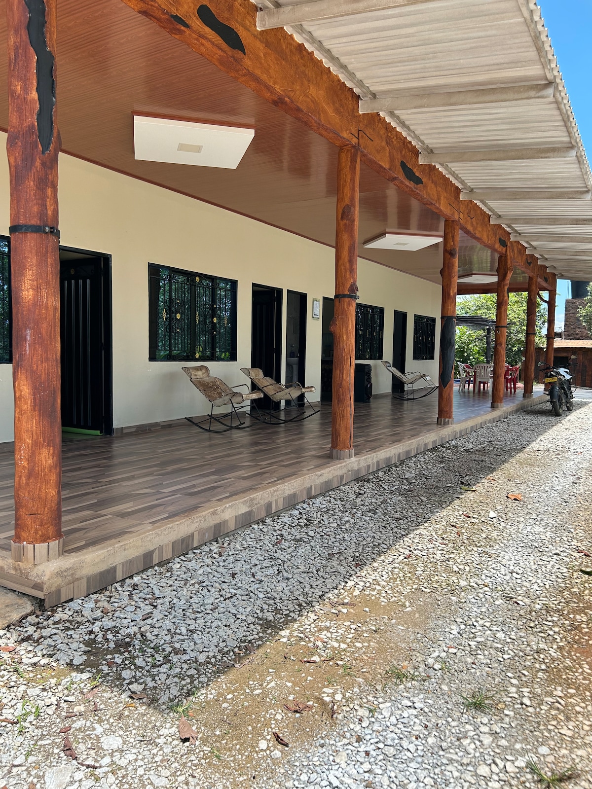A wide veranda features wooden beams and comfortably arranged lounge chairs. The flooring is covered in patterned tiles, leading to multiple doorways. Surrounding greenery can be seen beyond the porch, enhancing the sense of serenity.