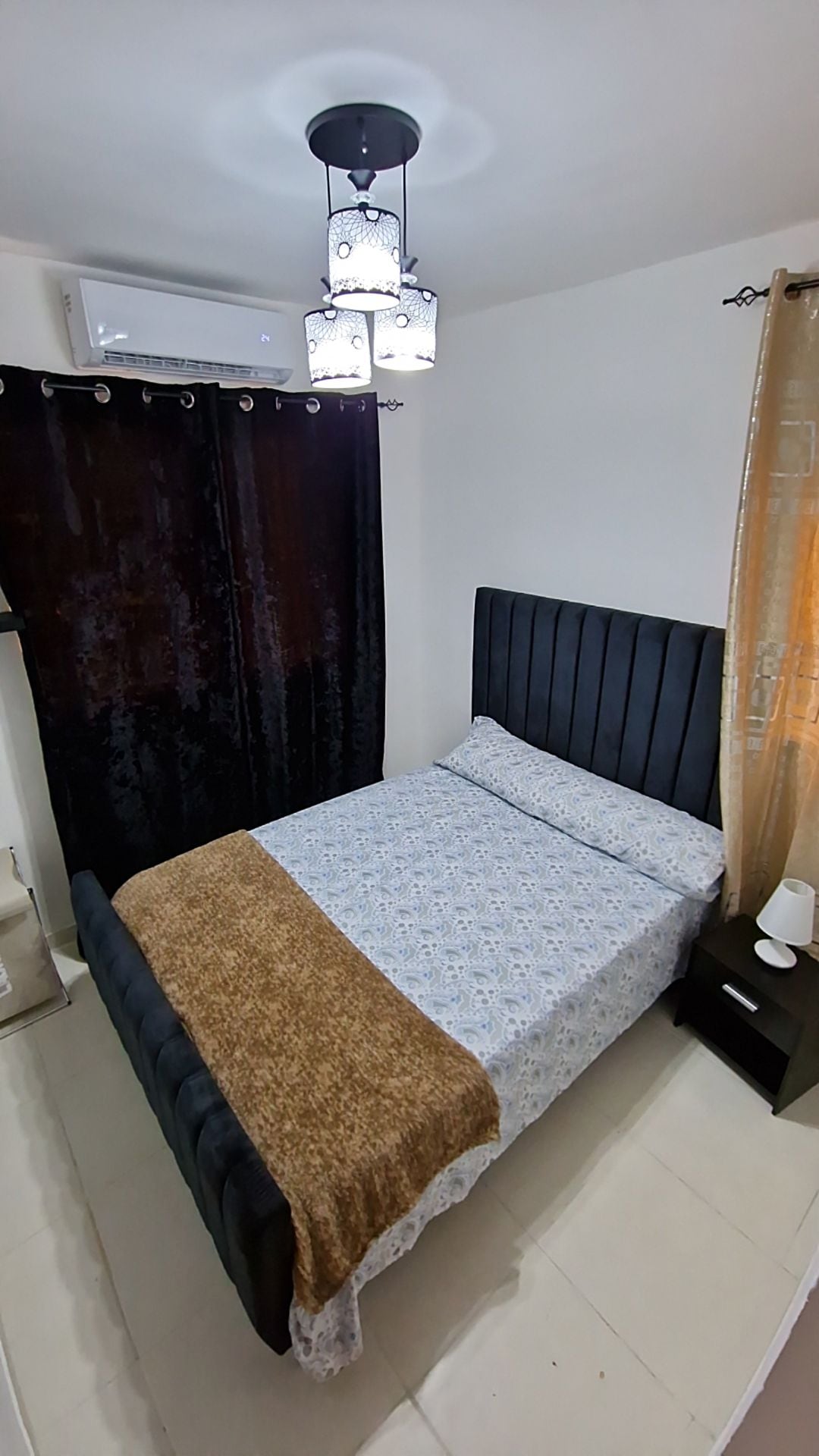 A cozy sleeping area is highlighted by a stylish bed with a dark headboard and a decorative throw blanket. Soft lighting from a unique ceiling fixture enhances the serene atmosphere. The room features black curtains alongside light beige drapes, creating a balanced contrast.