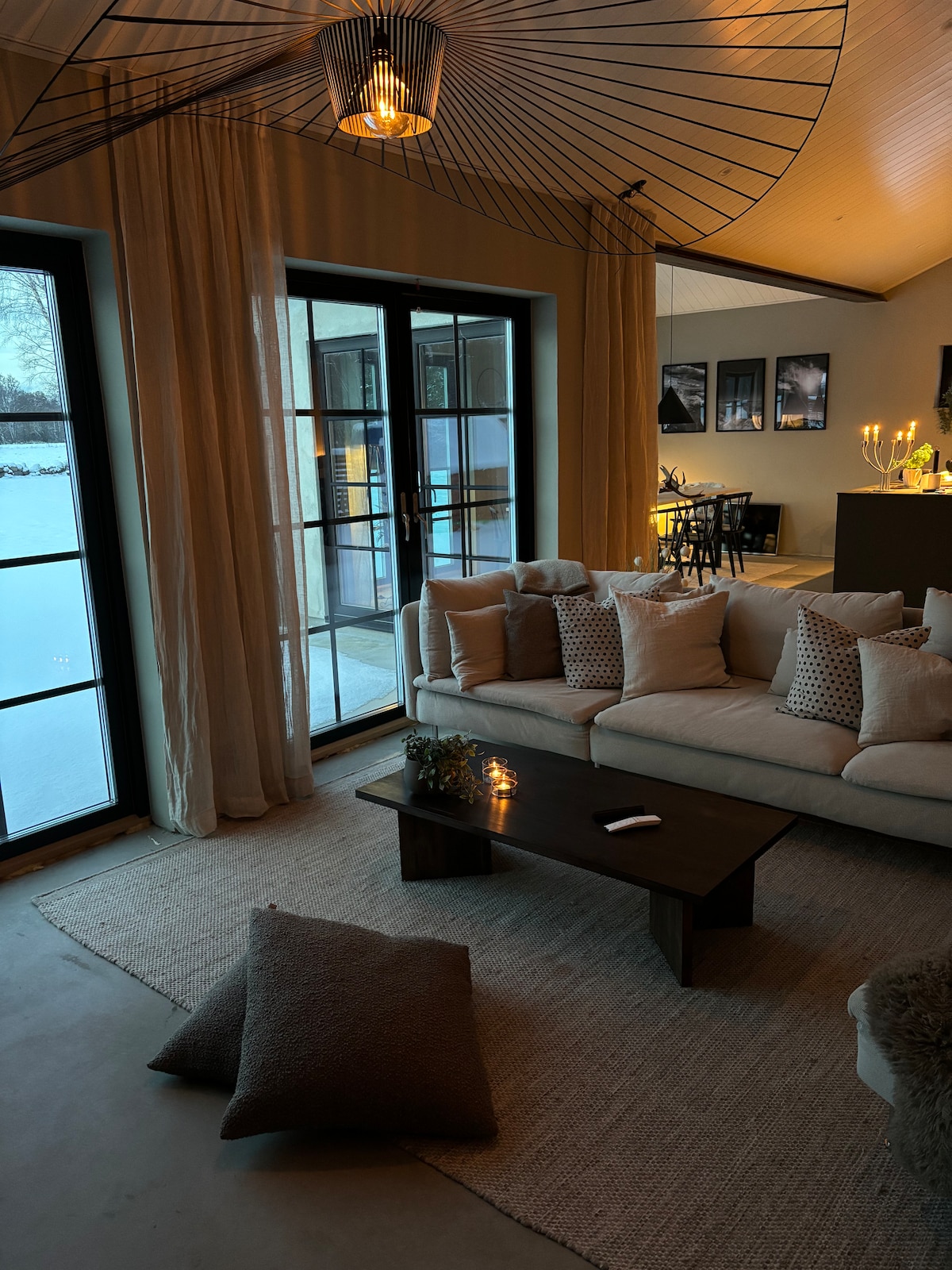 A spacious living area features large glass doors that open to a snowy landscape. Soft, neutral-toned cushions adorn the sofa. A textured rug lies on the floor, accompanied by decorative pillows. Warm lighting is provided by an overhead fixture, creating a cozy ambiance.