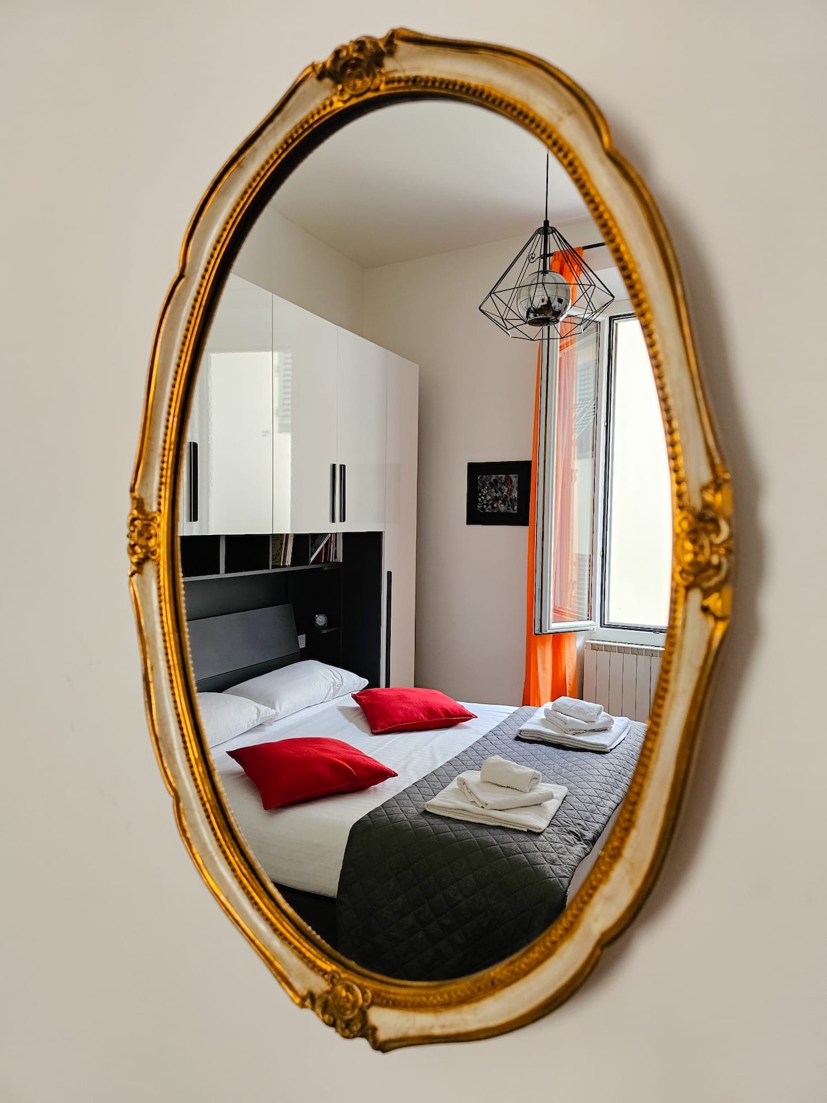 A vintage oval mirror frames a cozy bedroom. The bed is adorned with red accent pillows and a patterned blanket. A window with orange drapes allows natural light to brighten the space, while a geometric pendant light hangs above the bed.