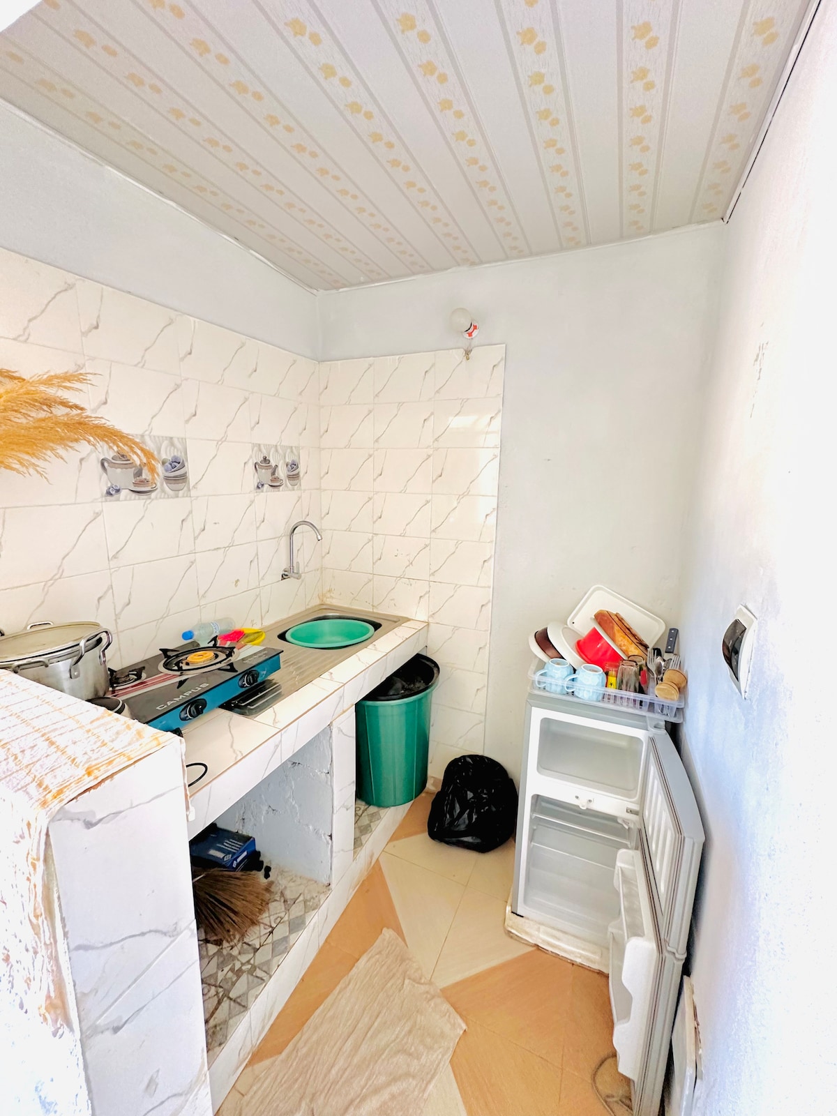 A compact kitchen area features a marble countertop with a sink and stovetop, with various kitchen utensils arranged nearby. A small fridge is visible to one side, while a green trash bin and a storage box are positioned against the wall.