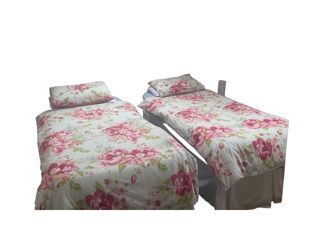 The image features two single beds with floral-patterned bedding in soft pastel colors. Each bed is adorned with matching pillows and a comfortable mattress. A bedside table is visible near one of the beds, providing a convenient surface for personal items.
