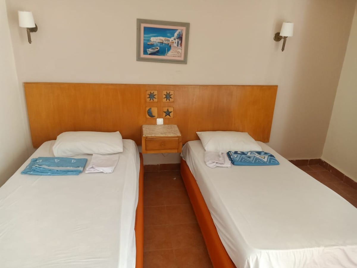 A simple sleeping area features two separate beds with white linens, each accompanied by a blue towel. A wooden headboard adds warmth to the space, while a small nightstand is positioned between the beds. Distinctive artwork adorns the wall above the nightstand.
