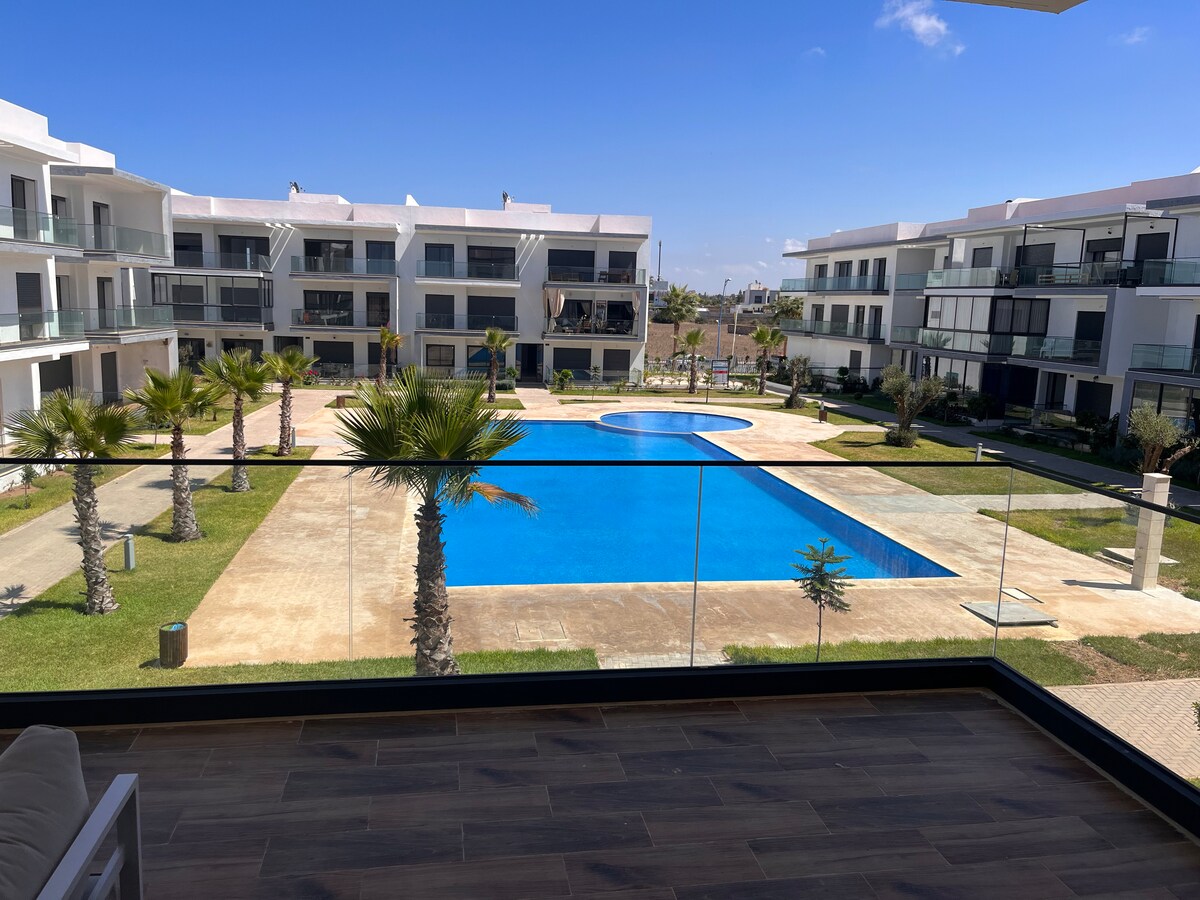 A spacious terrace offers a clear view of a large swimming pool surrounded by well-maintained gardens and palm trees. Modern residential buildings are visible in the background against a bright blue sky.