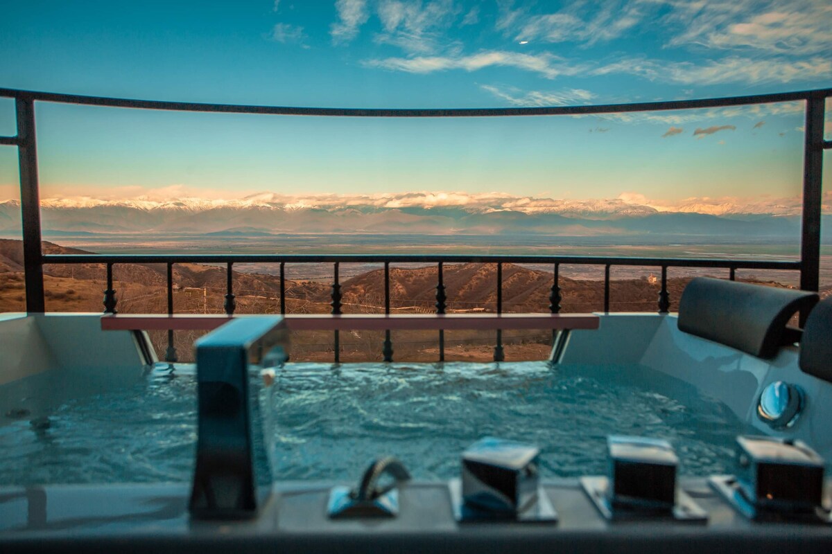 A jacuzzi on a terrace offers panoramic views of distant mountains under a clear blue sky. The water's surface reflects the surrounding landscape, while the railing provides a safe enclosure, enhancing the outdoor relaxation space.