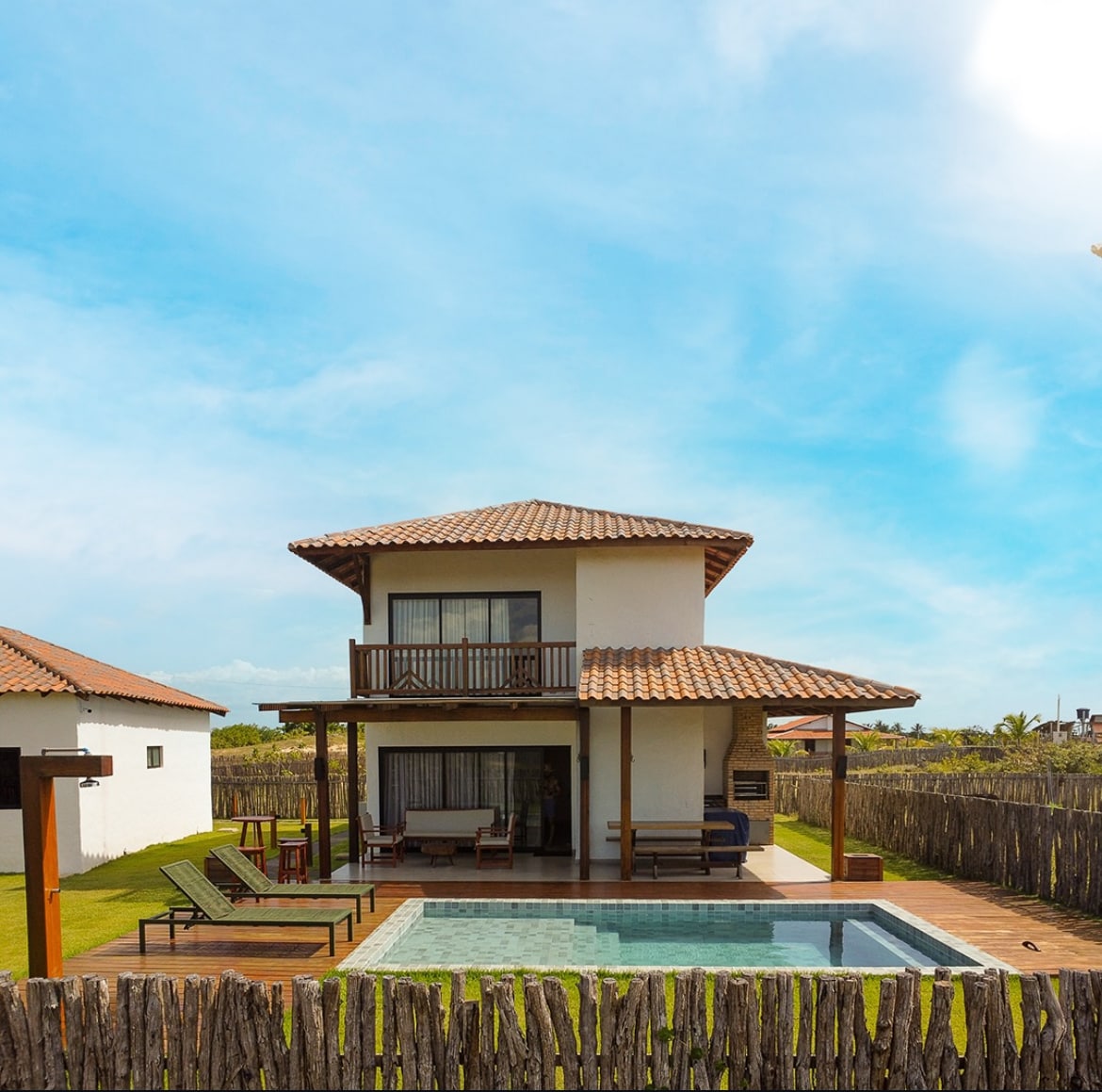A two-story house is set against a clear blue sky, featuring a charming tiled roof. A private pool is positioned in front, flanked by lounge chairs and a shaded seating area. Surrounding wooden fencing creates a sense of seclusion in the outdoor space.