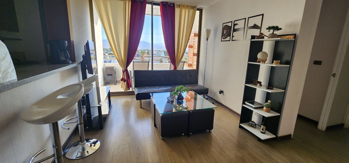A modern living area features a sleek black sofa opposite a glass coffee table. Large windows draped with colorful curtains allow natural light to fill the space. A slender bookshelf holds decorative items, while a bar with high stools is positioned near the kitchen area.