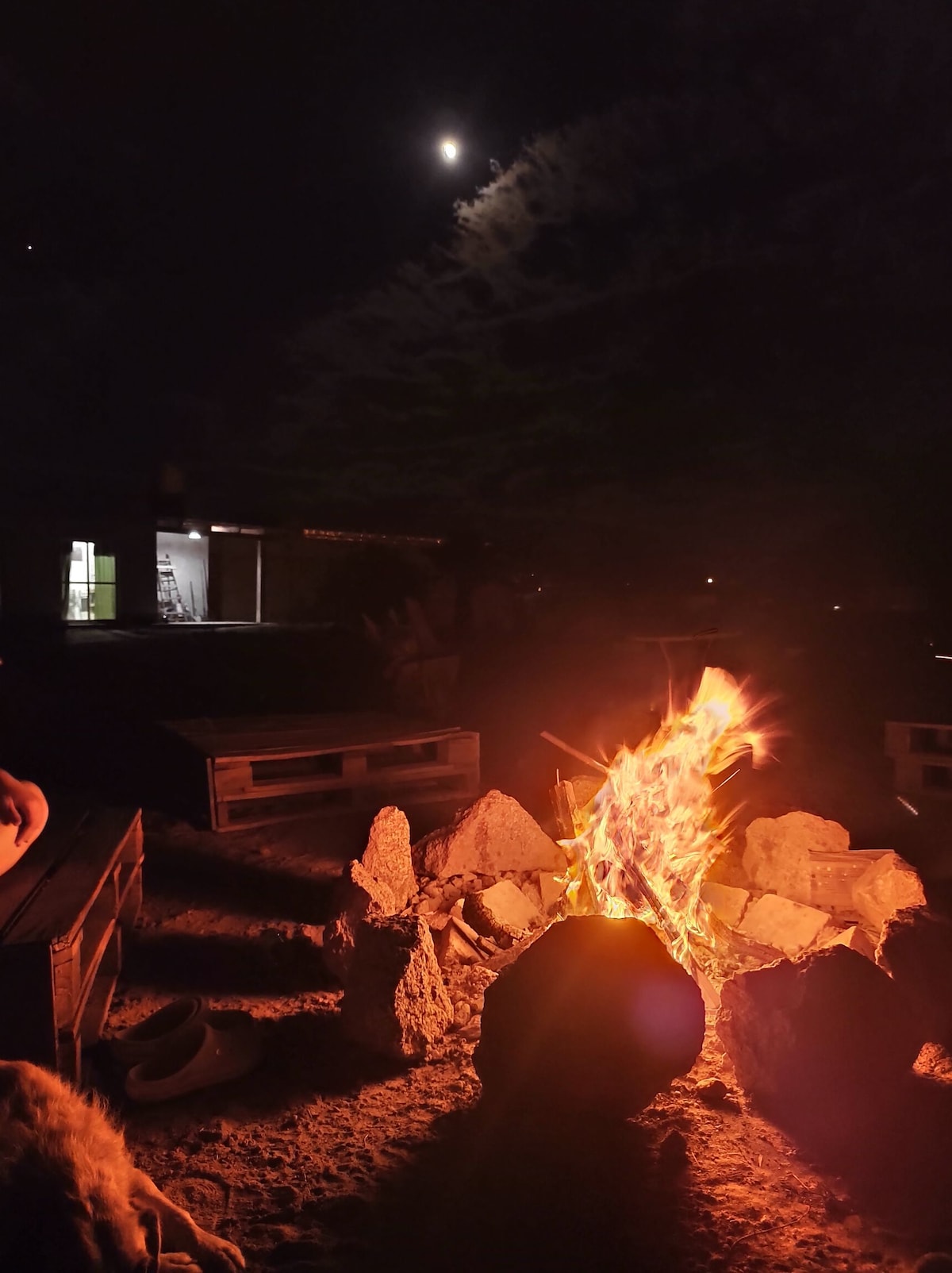 A warm campfire glows brightly in the foreground, surrounded by rocks. The night sky is illuminated by the moon and scattered clouds, creating a serene atmosphere. A distant structure is faintly visible in the background, suggesting nearby accommodations.