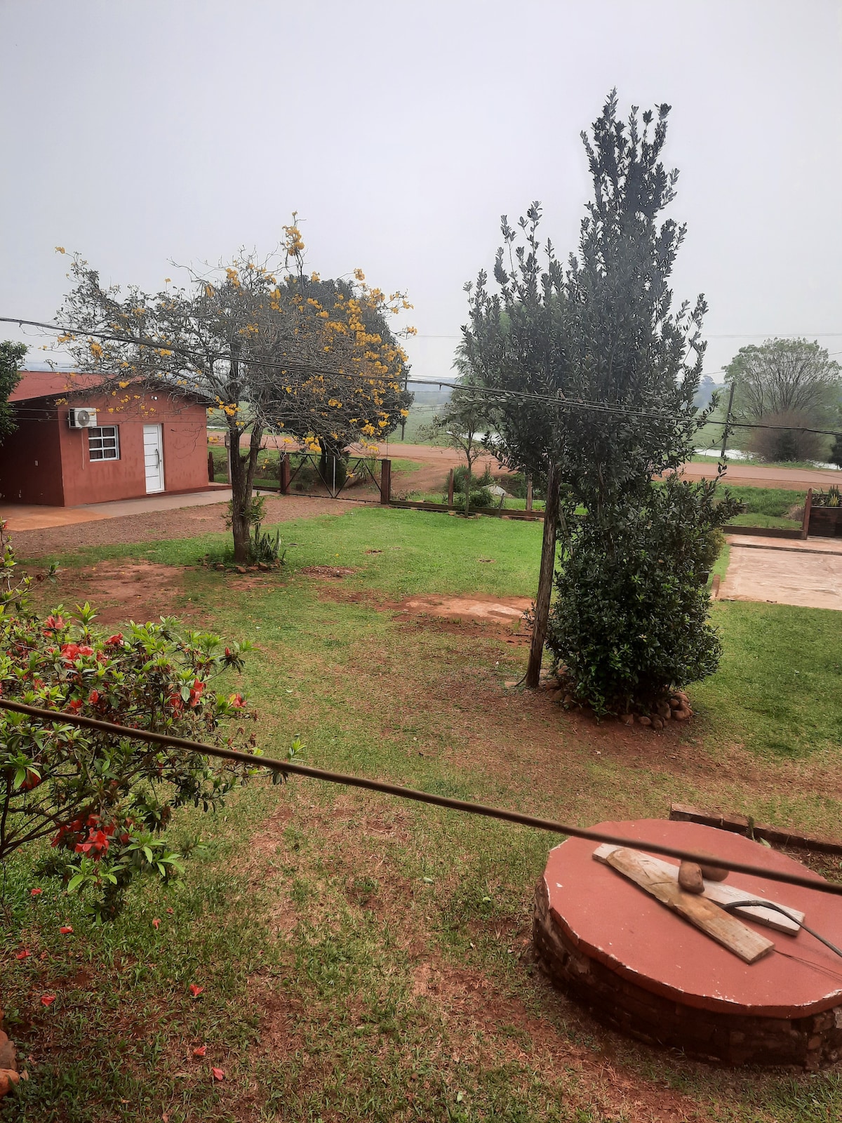 A green outdoor space is showcased, featuring well-maintained grass and a variety of trees. A small red structure is visible in the background, complemented by a circular stone well in the foreground. The scene is serene and inviting, perfect for family gatherings.