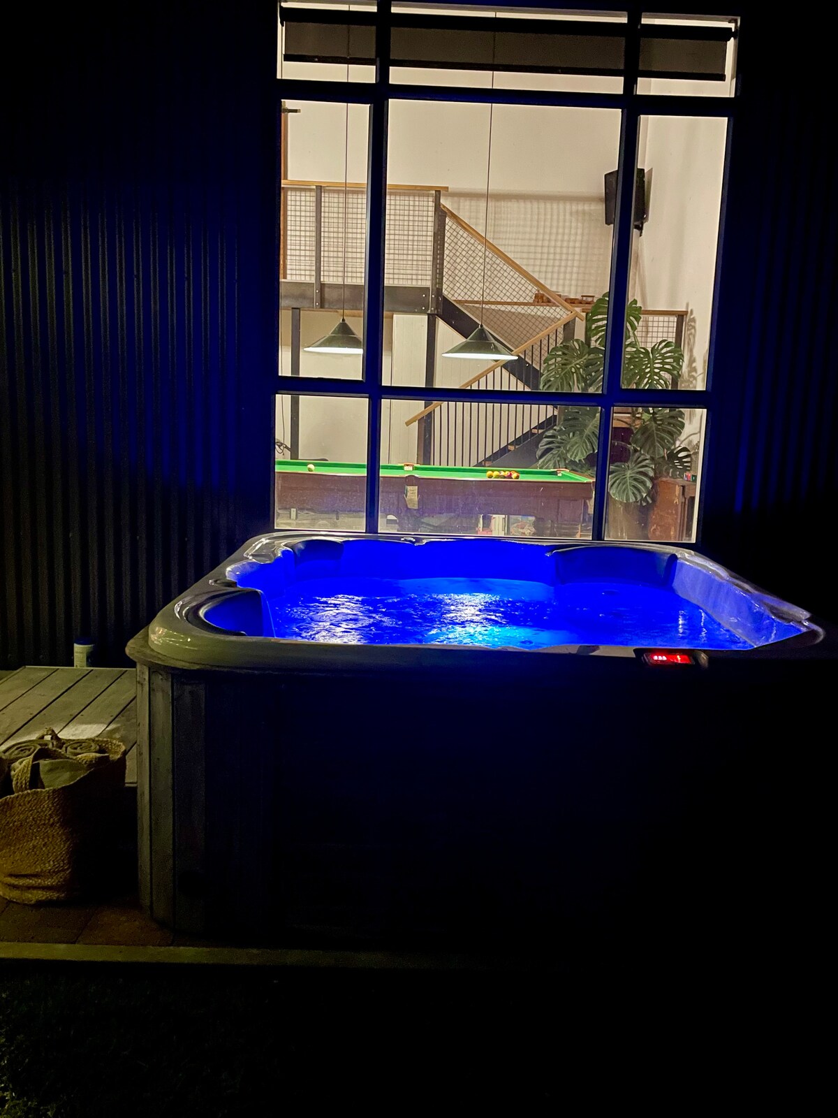 A spa pool is illuminated in blue light, providing a serene ambiance. A large window reveals an open space with a staircase and greenery in the background. Natural wood textures and soft lighting complement the cozy atmosphere.