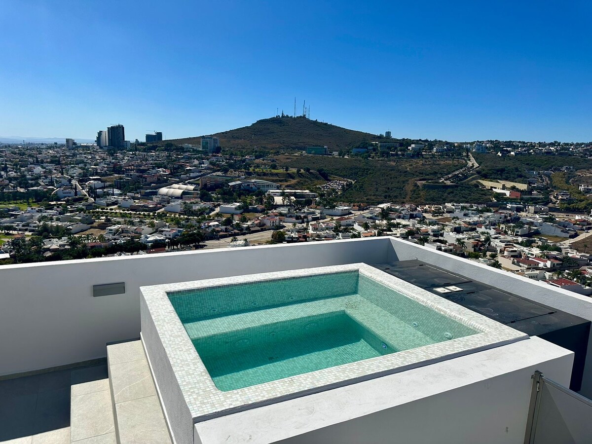 A private terrace features a rectangular jacuzzi surrounded by light-colored, smooth tiles. Expansive views of the city landscape and distant mountains are visible, under a clear blue sky. The setting highlights both relaxation and accessibility to outdoor space.