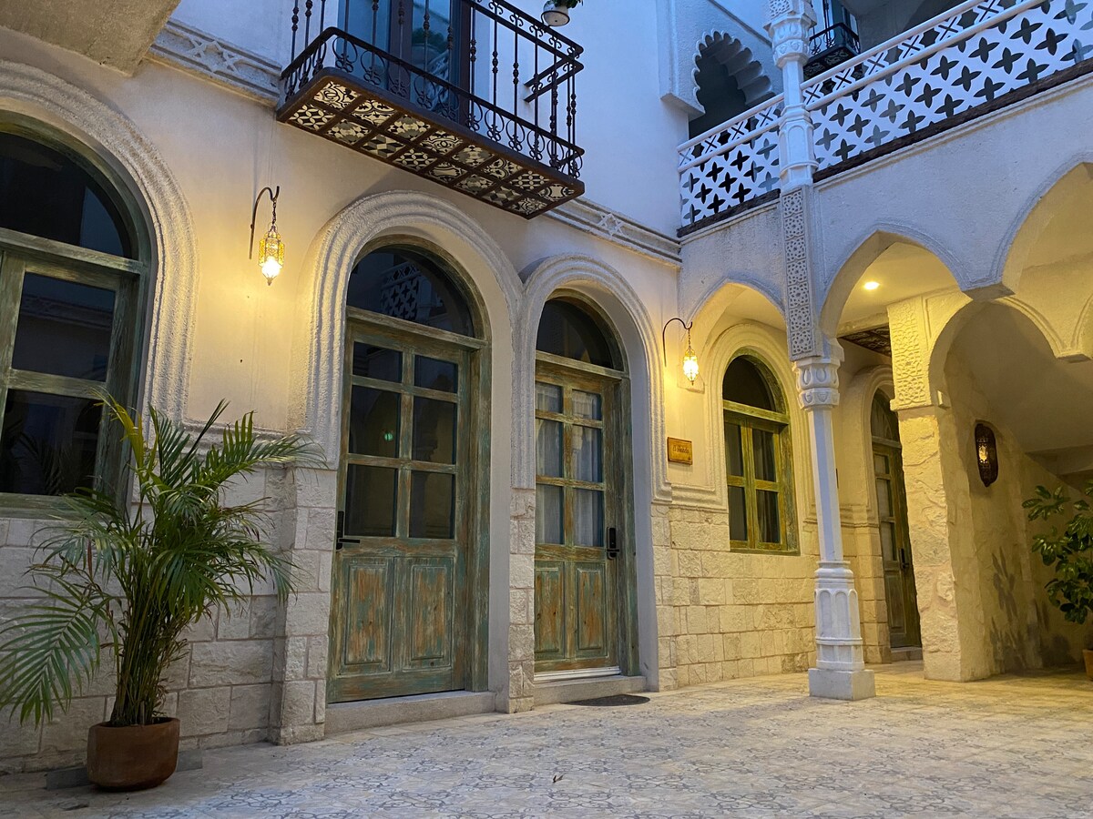 An elegant courtyard is shown, featuring two wooden doors with green accents set in a stone wall. Soft lighting illuminates the area, enhancing the intricate architectural details, while a potted plant adds a touch of nature to the scene.