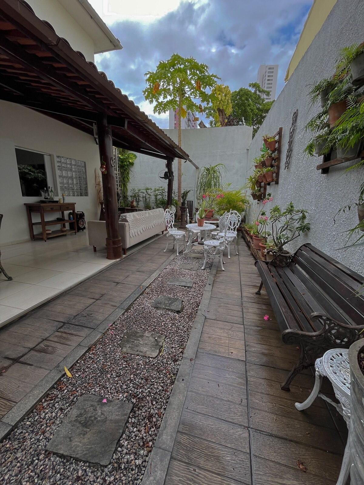An outdoor area with a well-maintained pathway made of stone and wood leads through a garden. White furniture with intricate designs is arranged for seating, while lush greenery and ornamental plants are positioned throughout the space, providing a relaxing ambiance.
