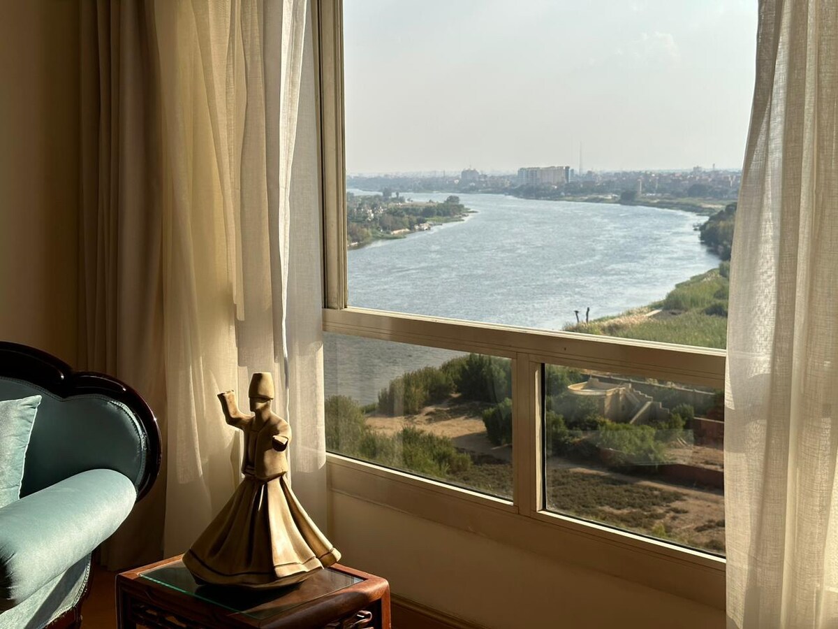 A serene view of a river is framed by large windows adorned with soft, sheer curtains. The focal point is a decorative figure on a wooden table, inviting tranquility while overlooking the lush greenery along the riverbank.