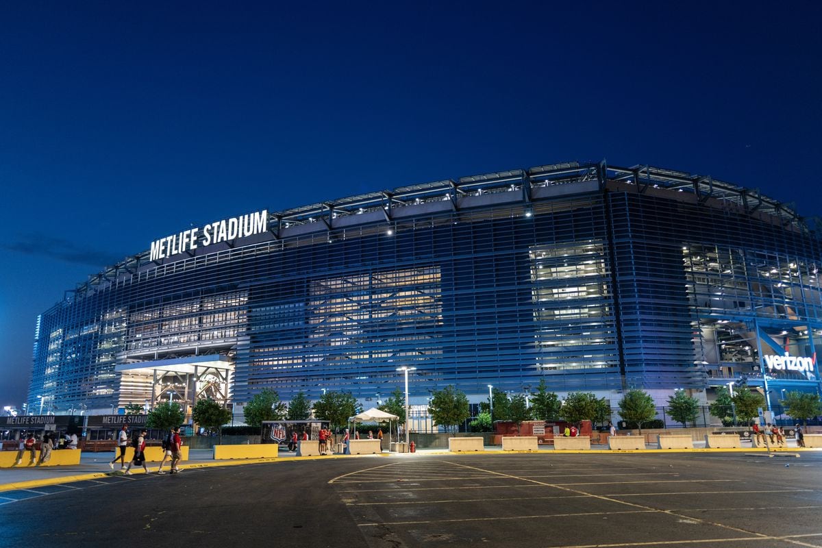 The exterior of MetLife Stadium is illuminated against a twilight sky, showcasing a modern architectural design. Adjacent trees line the foreground, while the stadium's name is prominently displayed. The scene captures the ambiance of an active event venue with distant attendees and vehicles parked nearby.