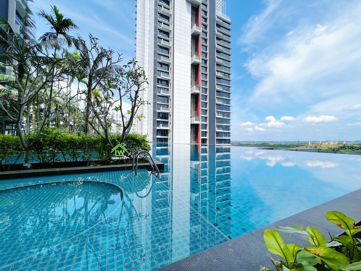 A serene infinity pool is framed by lush greenery, showcasing shimmering blue tiles that reflect the sky. Towering residential buildings rise in the background, while the pool area provides a calm environment for relaxation.