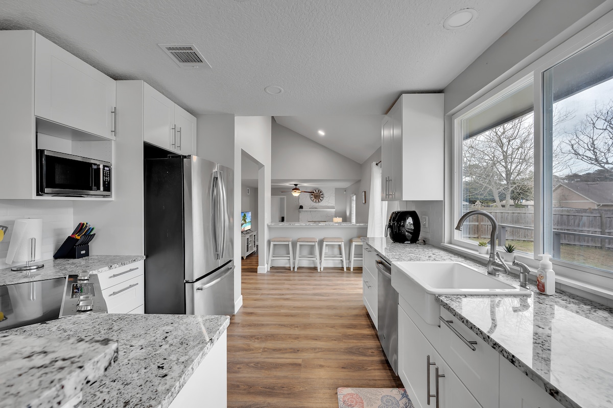 A modern kitchen features sleek white cabinetry and a spacious countertop made of light-colored granite. Stainless steel appliances, including a refrigerator and microwave, are included. Large windows provide natural light, and a view of the backyard is visible, enhancing the open layout towards the adjacent dining area.