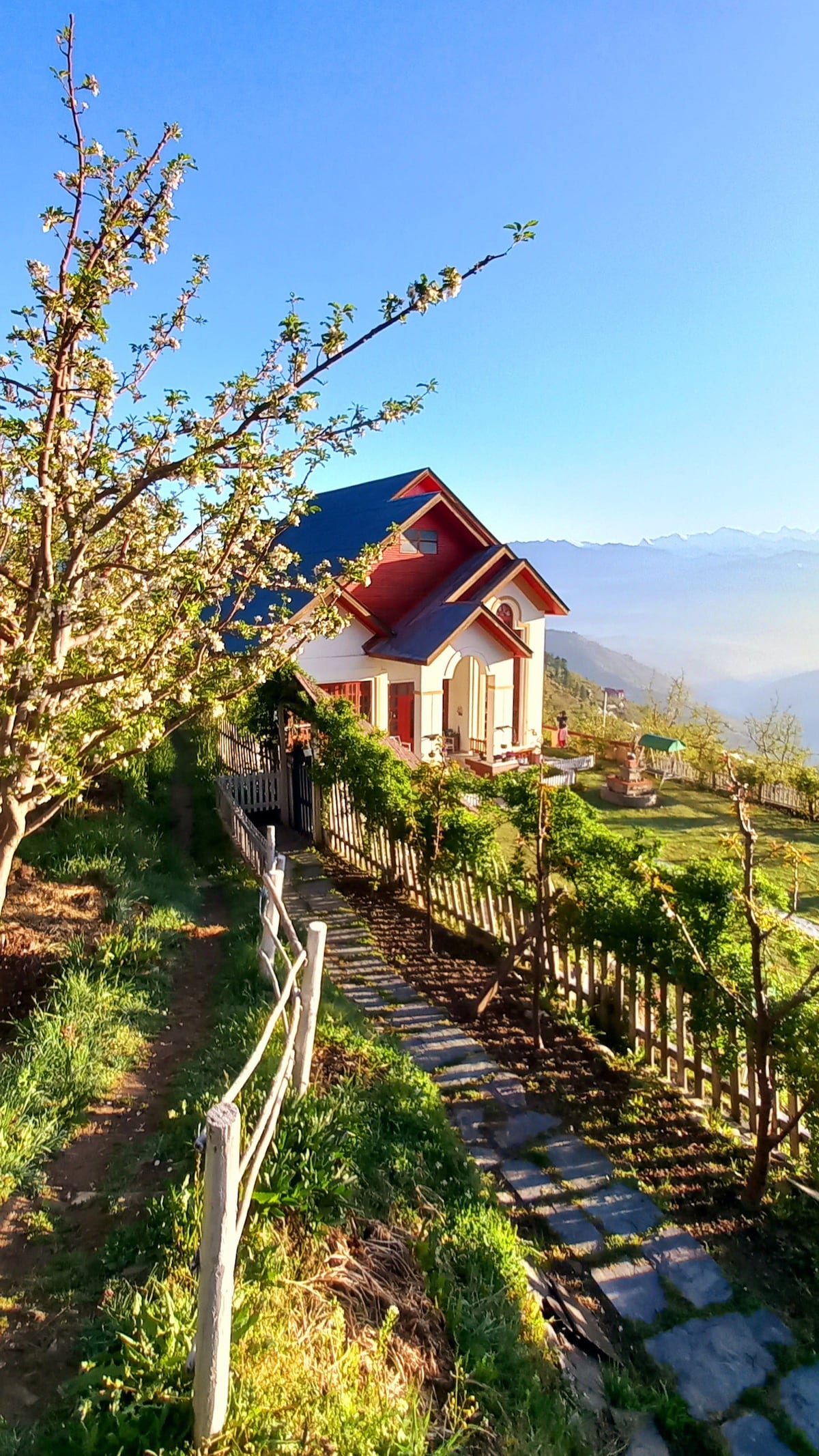A charming bungalow is nestled amidst lush greenery, featuring a vibrant red-roof and white walls. A winding stone path leads from a rustic wooden fence to the entrance, framed by blooming apple trees under a clear blue sky, with mountains visible in the distance.