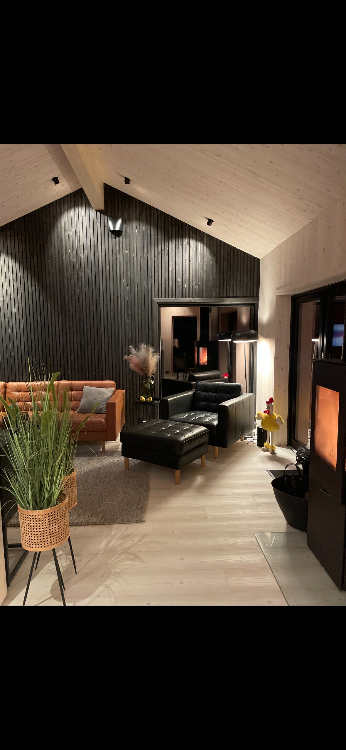 A modern living area features a mix of seating options, including a tan sofa and two black armchairs. Textured dark wood walls contrast with the lighter ceiling. Potted plants add a touch of greenery. Soft lighting creates a cozy ambiance, enhancing the space's inviting design.