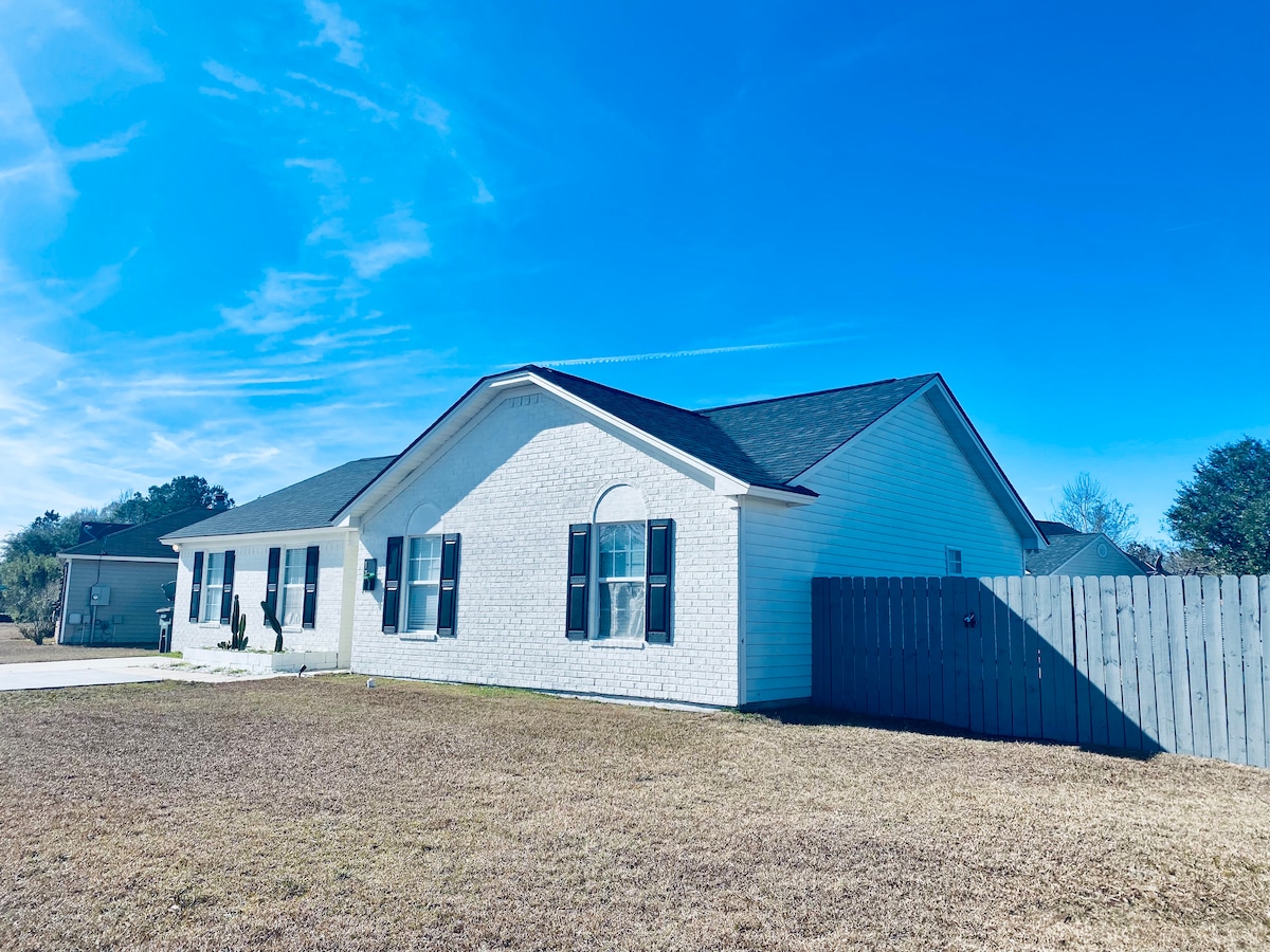The exterior of a single-story home is viewed, featuring a white brick facade and black shutters. A well-maintained front yard with grass is visible, alongside a grey wooden fence that encloses the property. A clear blue sky serves as the backdrop.