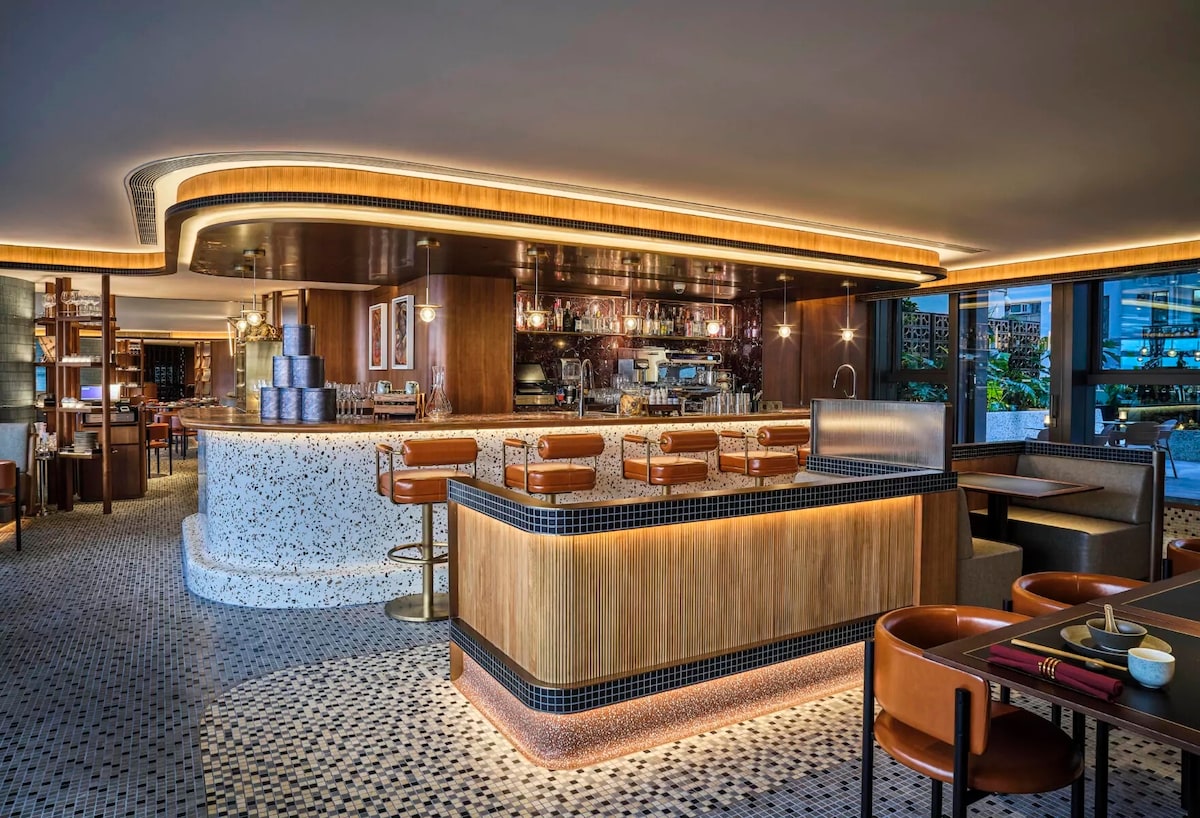 The image captures an elegantly designed bar area with a polished wooden counter and upholstered seating. Warm lighting accents the circular ceiling, and a variety of drink options are showcased behind the bar. A spacious dining area with modern furnishings is also visible.
